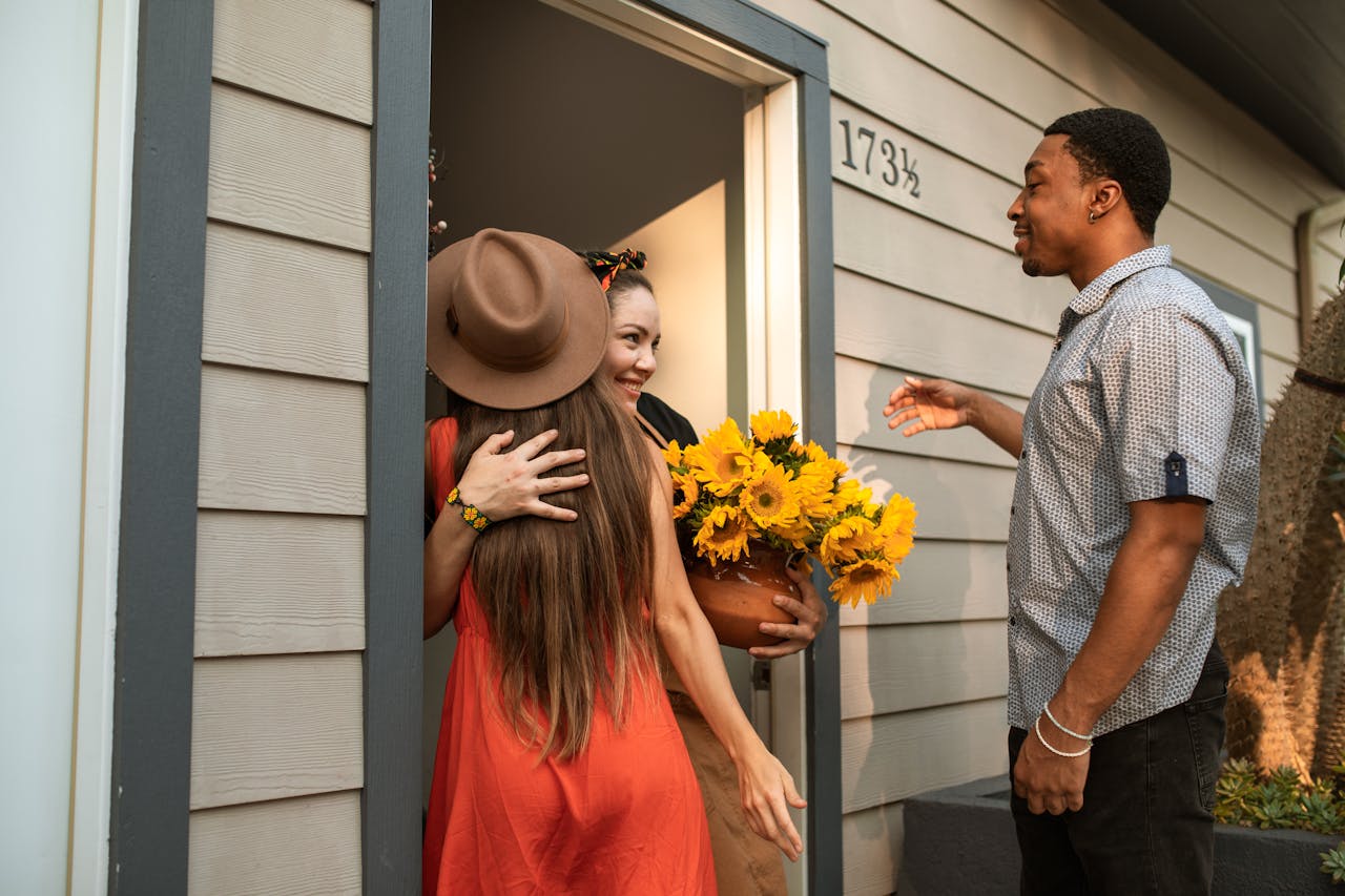 Three friends greeting and hugging at a house entrance with sunflowers.